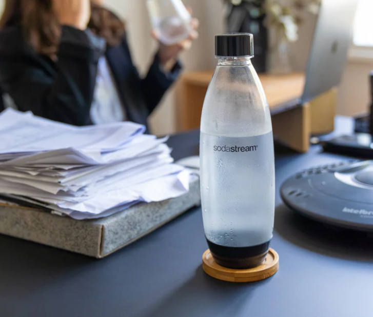 Eine klare Wasserflasche auf einem Holzuntersetzer. Schreibtisch mit Papieren, Laptop, Person unscharf.