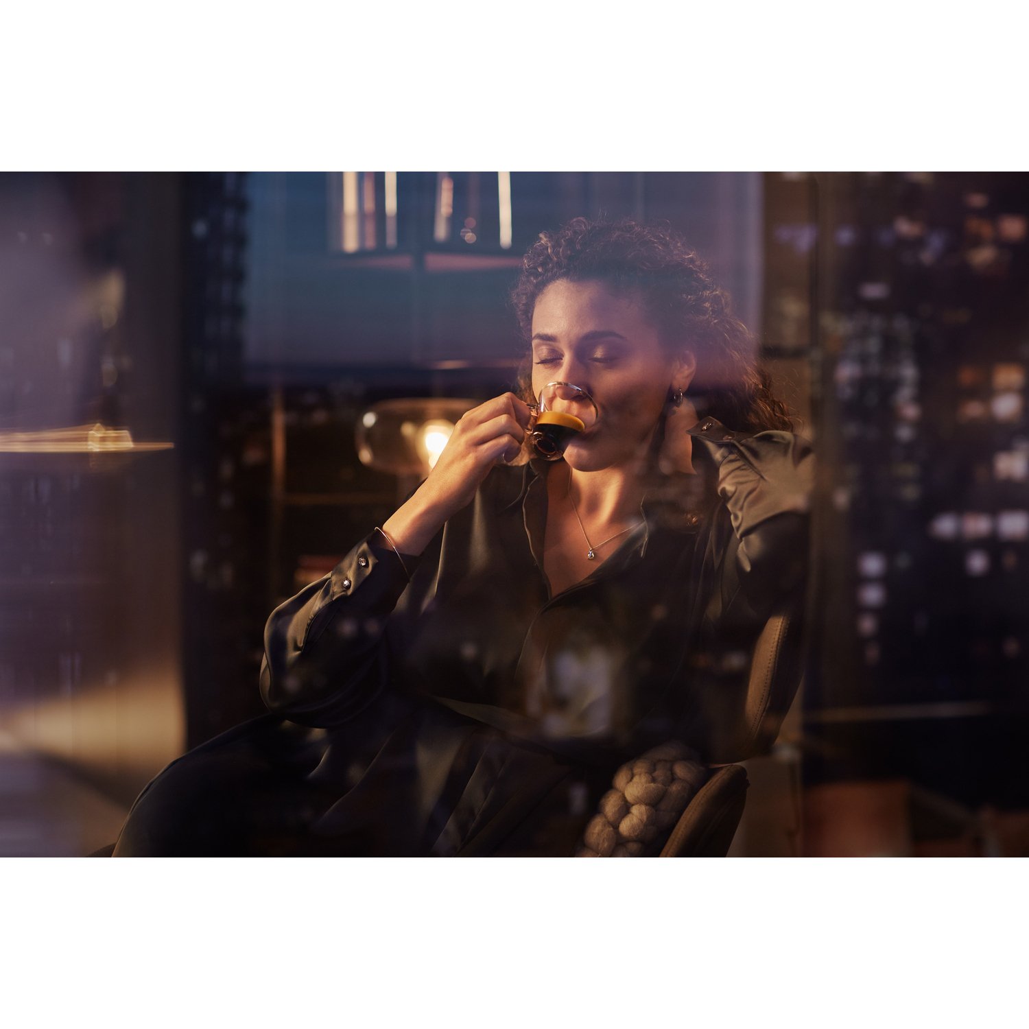 A woman is drinking coffee in a dark setting with a cityscape visible through the window.
