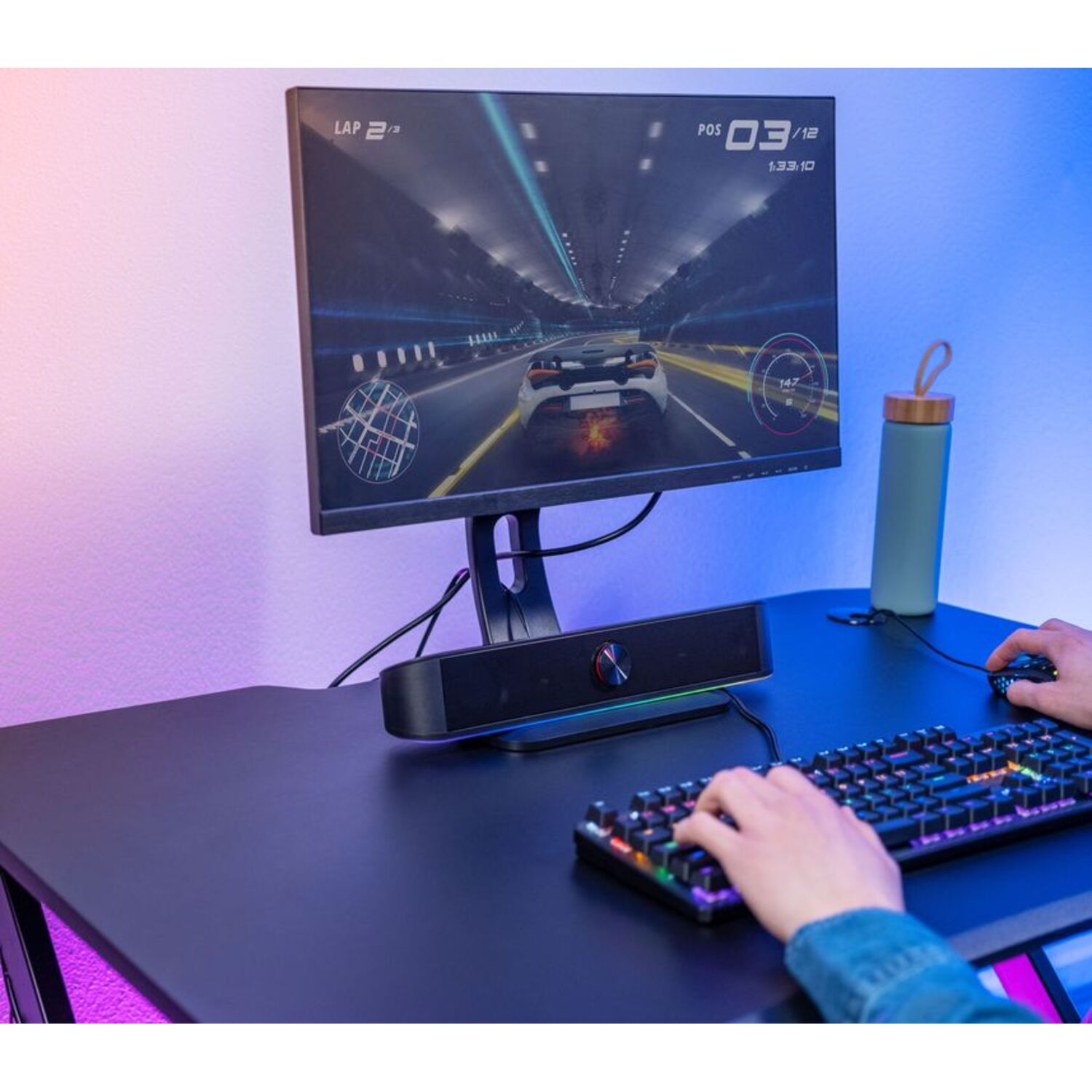 A gamer playing a racing game on a monitor, with a keyboard, speakers, and a water bottle.
