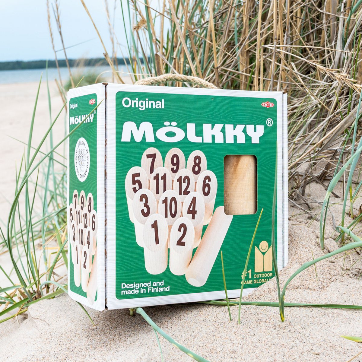 Zöld-fehér Molkky játékkarton homokos strandon, fűvel az előtérben és égbolttal a háttérben.