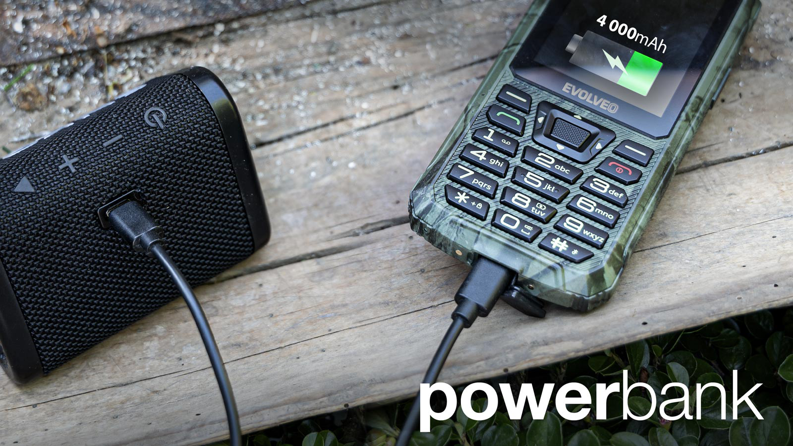 Egy fekete hangszóró, amely egy robusztus zöld telefont tölt fa deszkákon. Szöveg: powerbank.