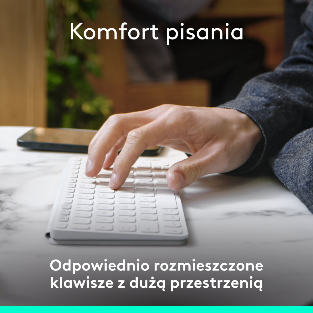 Dłoń pisze na białej klawiaturze. Tło obejmuje marmurowy stół, telefon i osobę w dżinsowej kurtce.