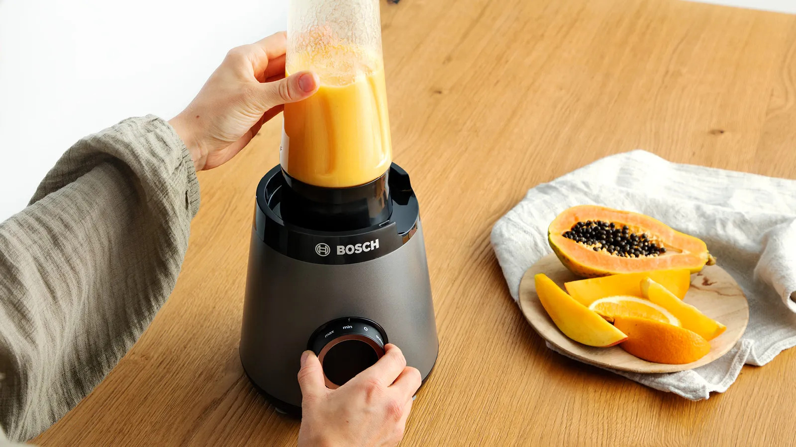 Osoba używa blendera z pomarańczowym płynem. Na drewnianym stole kawałki papai i pomarańczy.
