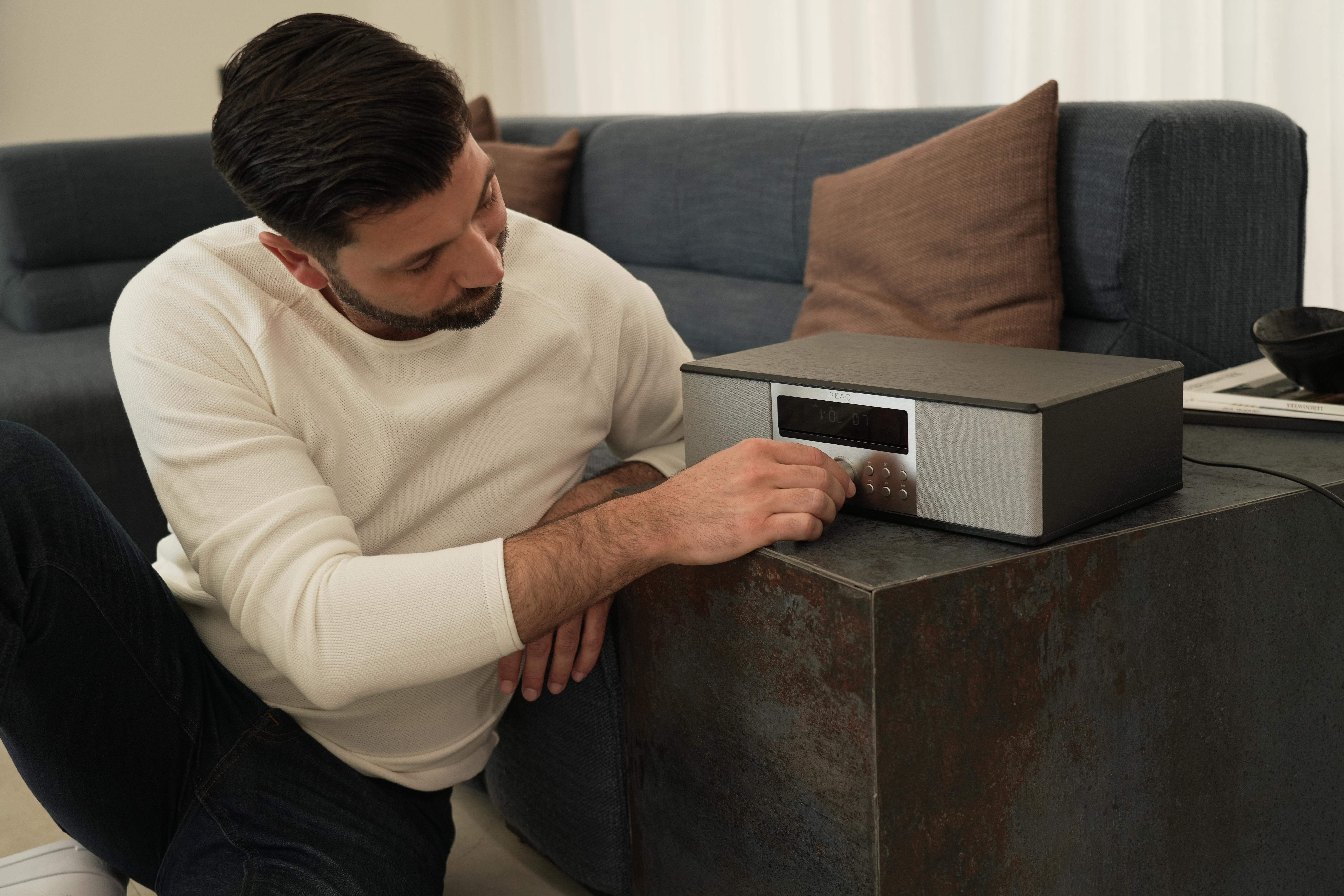 Ein Mann stellt einen Regler an einem Stereo ein, das auf einem Tisch neben einem blauen Sofa steht.