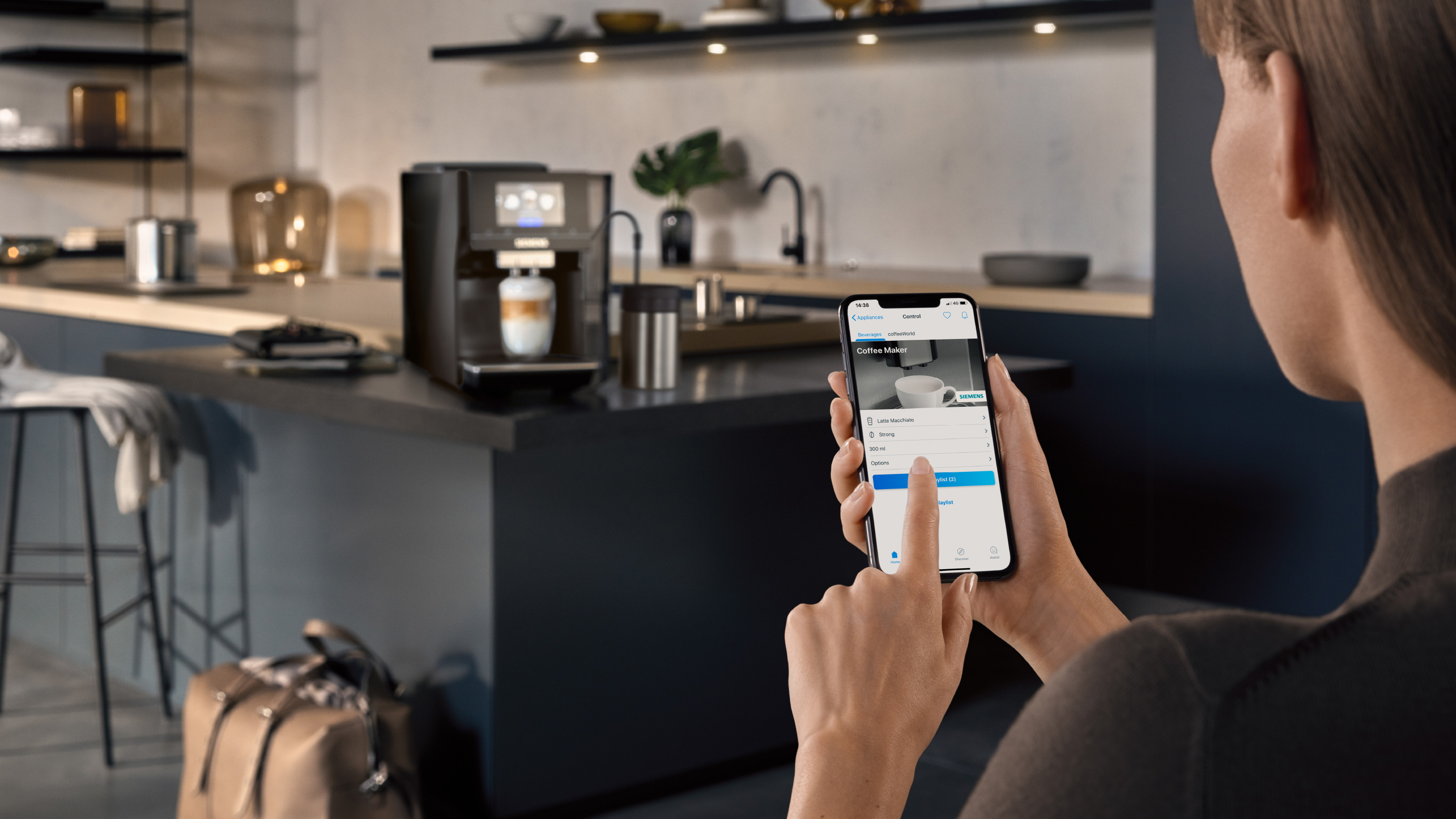 Frau bedient Kaffeemaschine per Handy. Küche im Hintergrund.