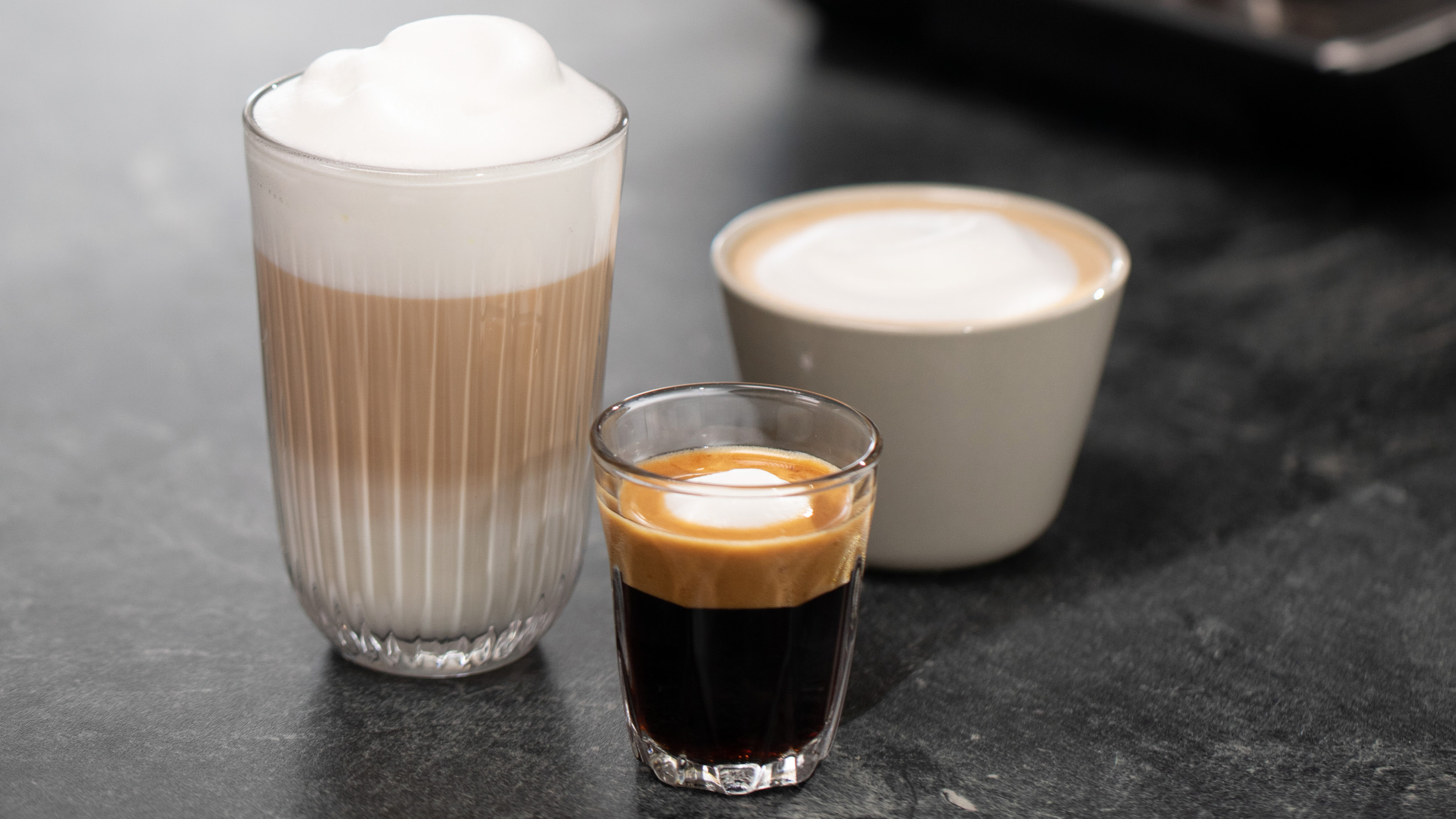 Drei Kaffeegetränke in Gläsern: Latte, Cappuccino und Espresso auf dunkler Oberfläche.