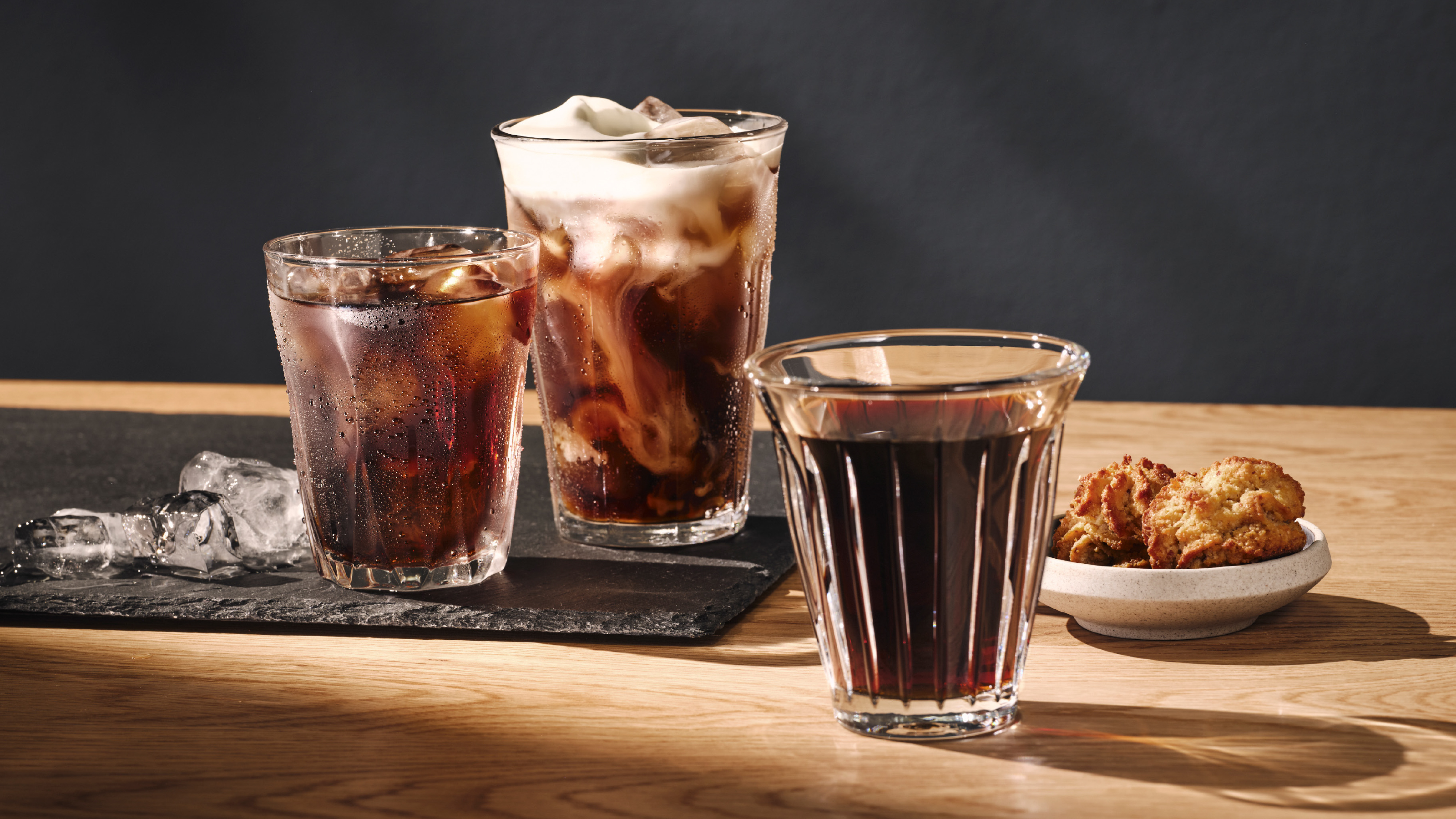 Drei Eiskaffeegetränke in Gläsern auf einem Holztisch mit dunklem Hintergrund.