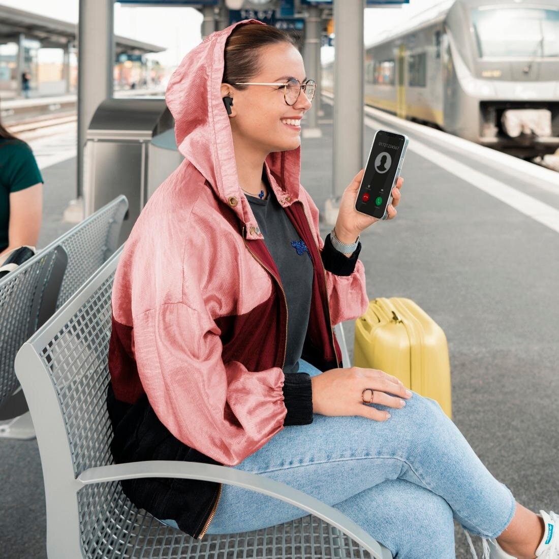 Kobieta na peronie rozmawia przez telefon. Ma na sobie różową kurtkę. W tle pociąg.
