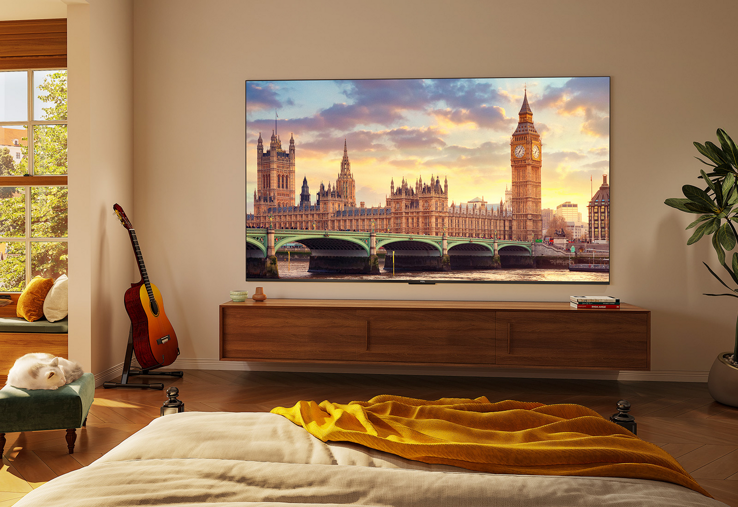 Sala de estar con un televisor grande que muestra una escena de Londres. Guitarra, cama, sofá con manta amarilla y plantas.