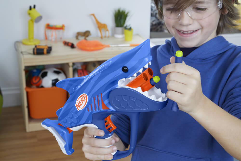 Junge hält eine blaue, haiförmige Spielzeugpistole. Er lächelt. Der Hintergrund zeigt Spielzeug.