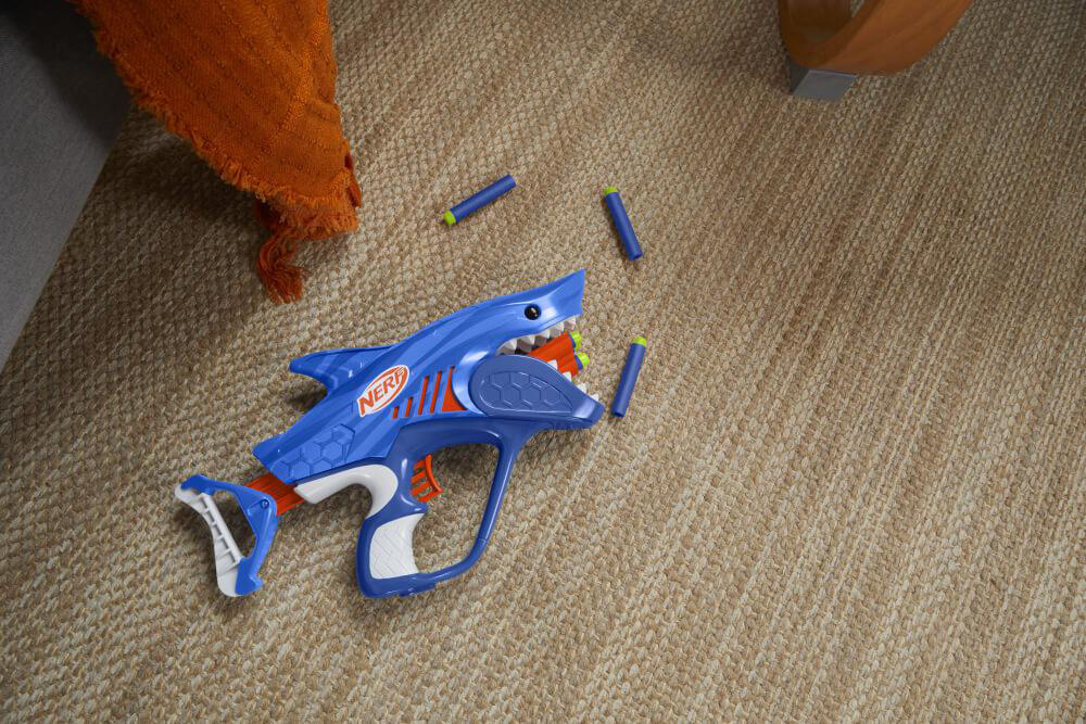 Blaue Hai-Nerf-Pistole auf braunem Teppich, mit orangefarbenem Handtuch.