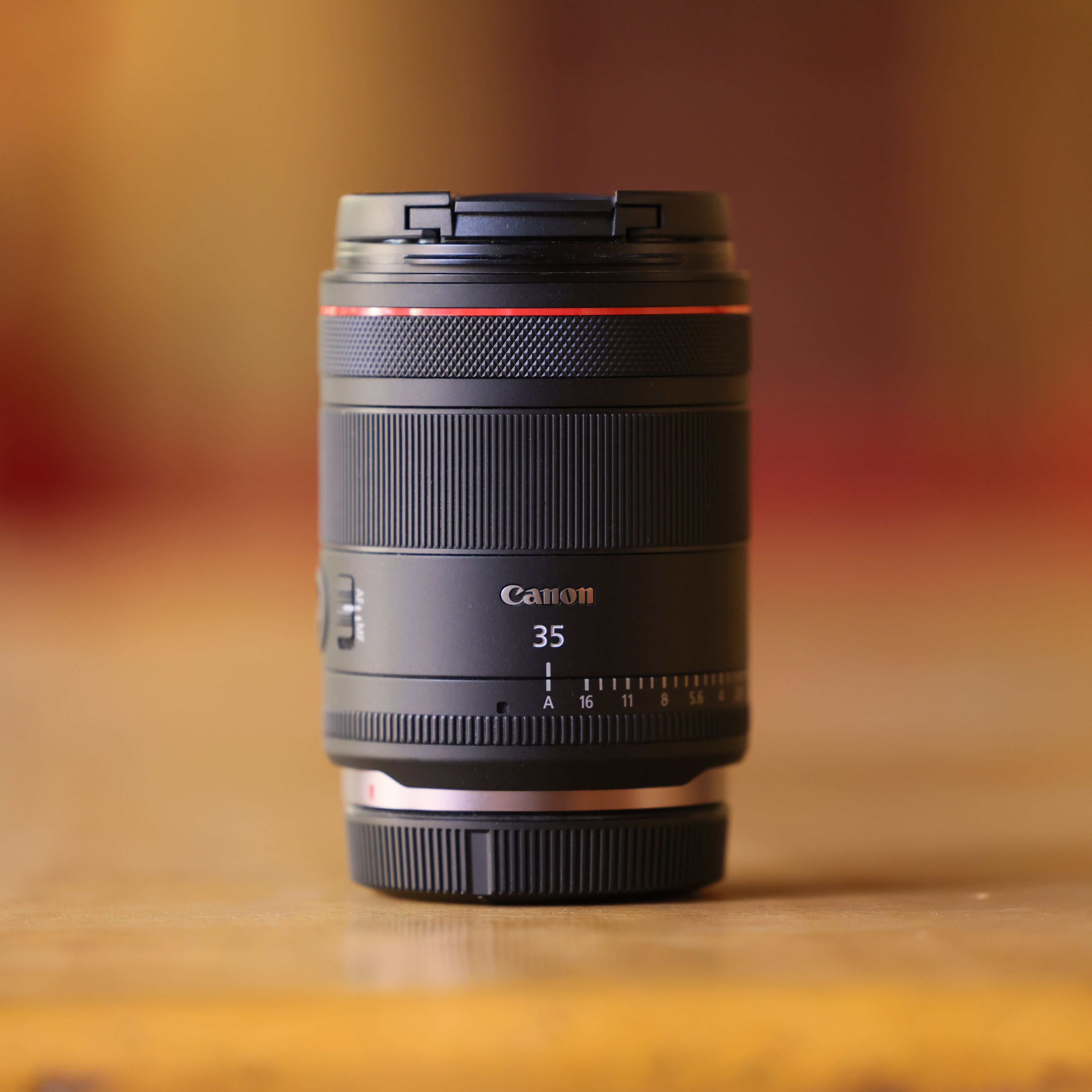 Objectif Canon 35mm noir avec anneau rouge, sur bois. Le fond est un dégradé flou orange et marron.