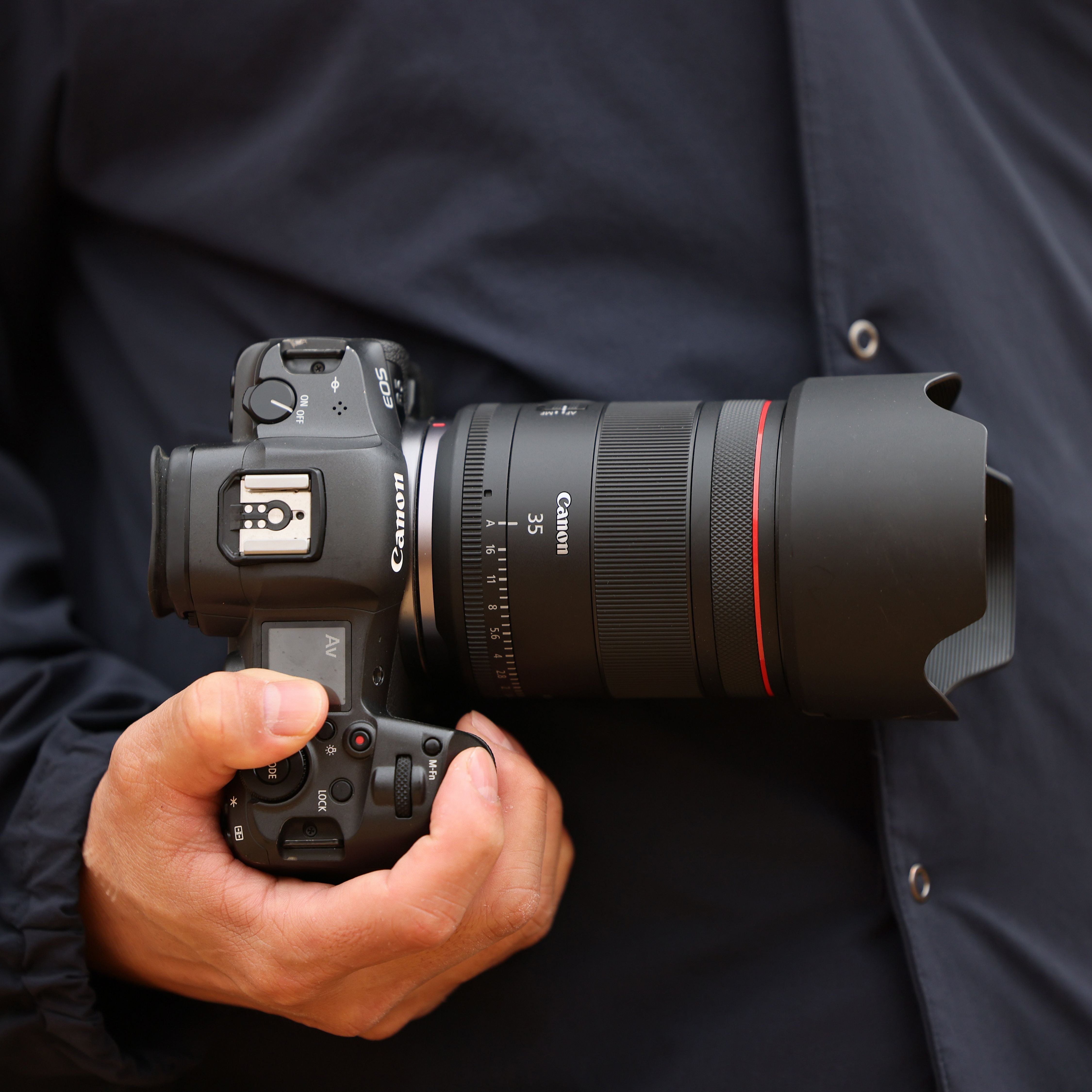 Une personne tient un appareil photo Canon avec un objectif. L'appareil photo est noir. Le fond est une veste sombre.