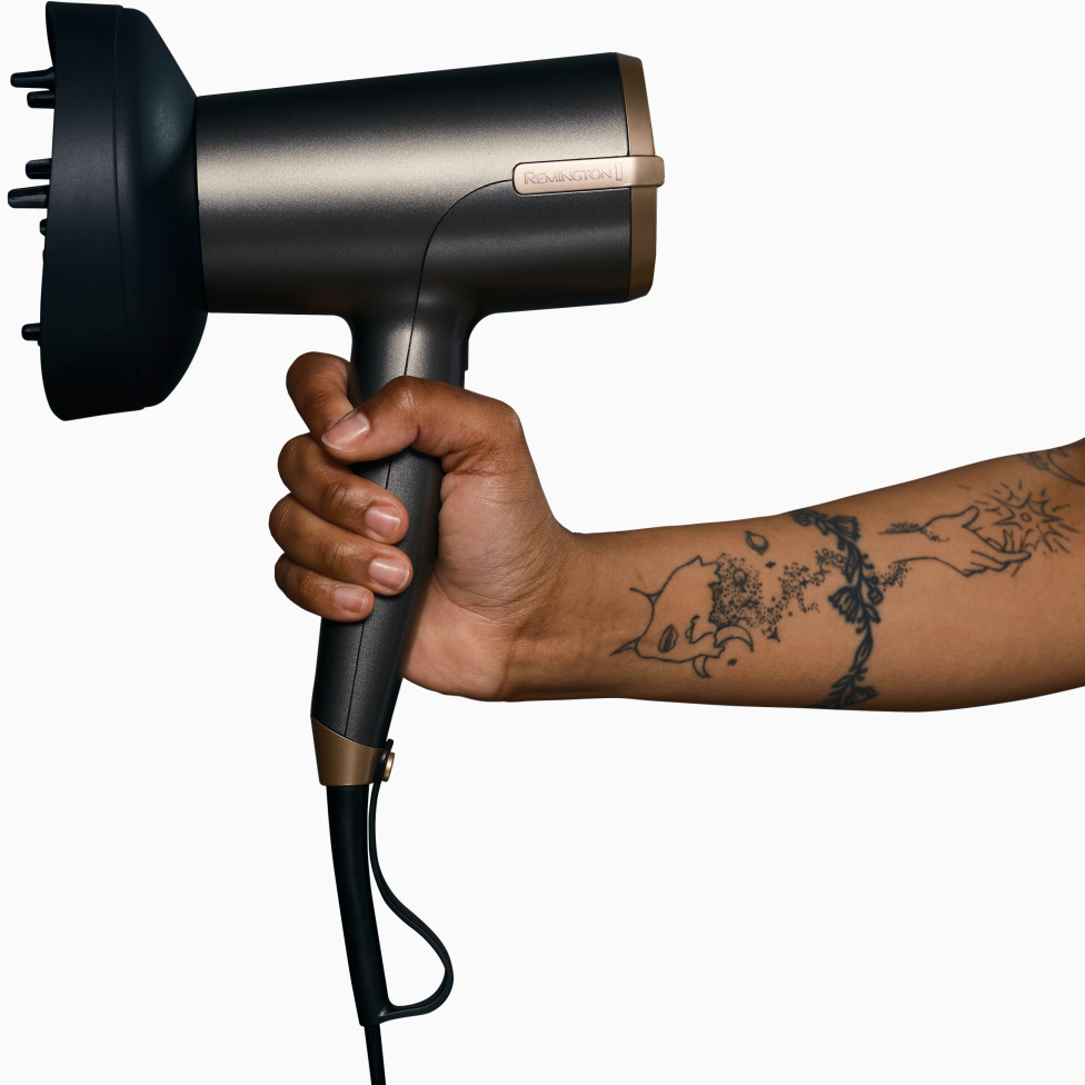 Persona tiene asciugacapelli nero e oro con tatuaggi sul braccio, sfondo bianco.