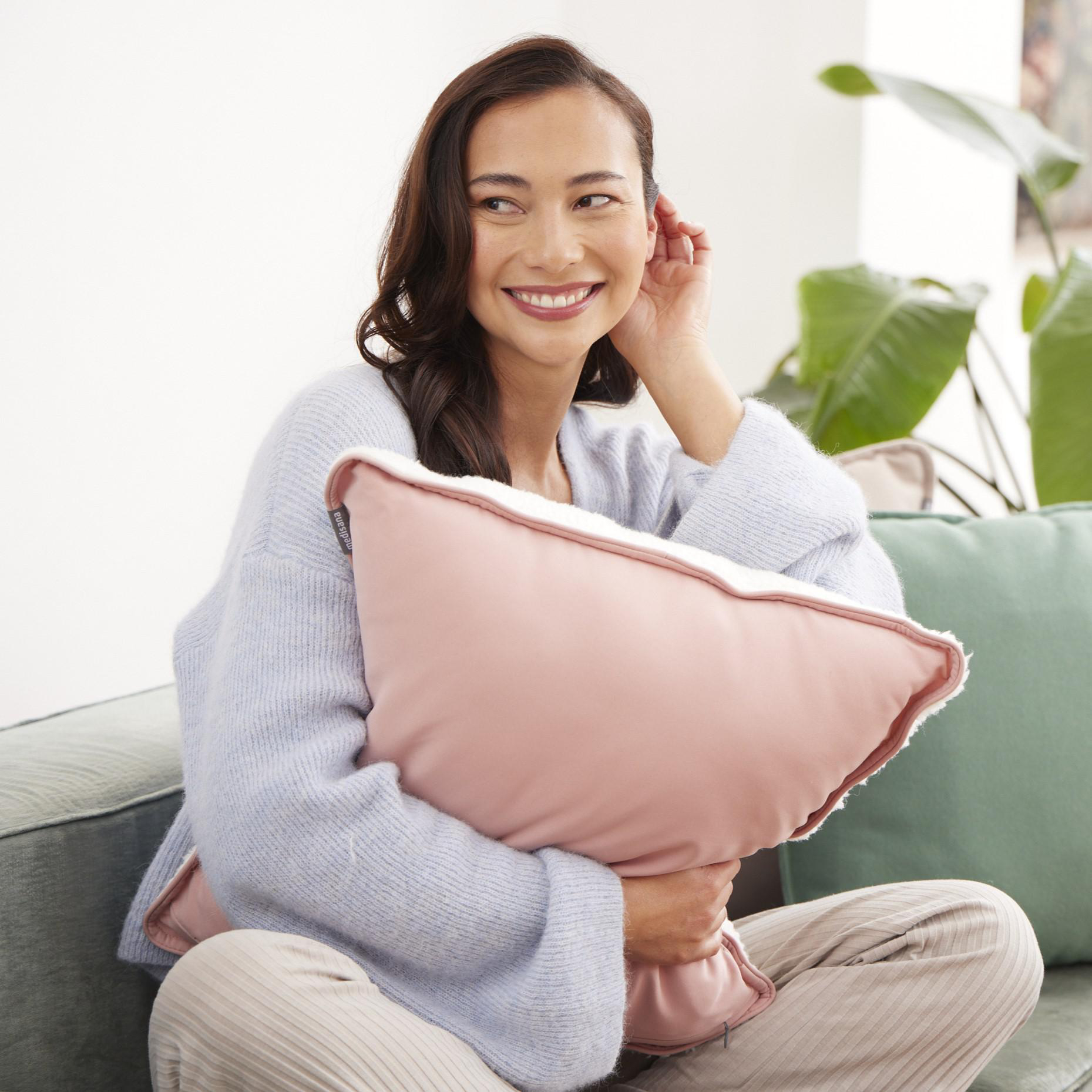 Eine Frau mit pinkfarbenem Kissen auf einer Couch, lächelnd. Grüne Pflanze im Hintergrund.