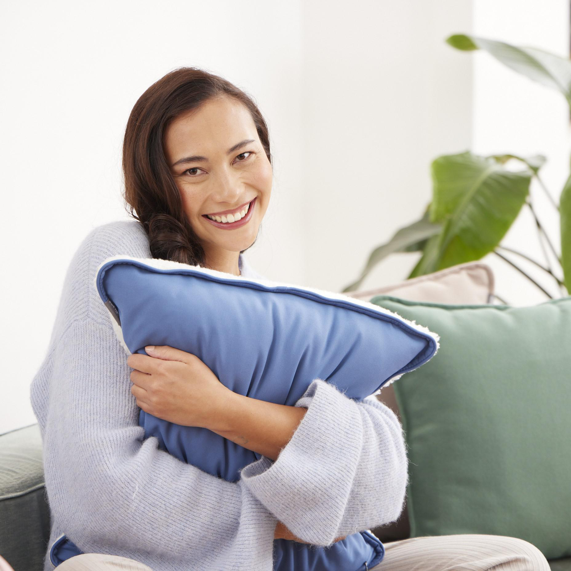 Frau hält blaues Kissen, lächelt. Grüne und beige Kissen, Sofa und Pflanze im Hintergrund.
