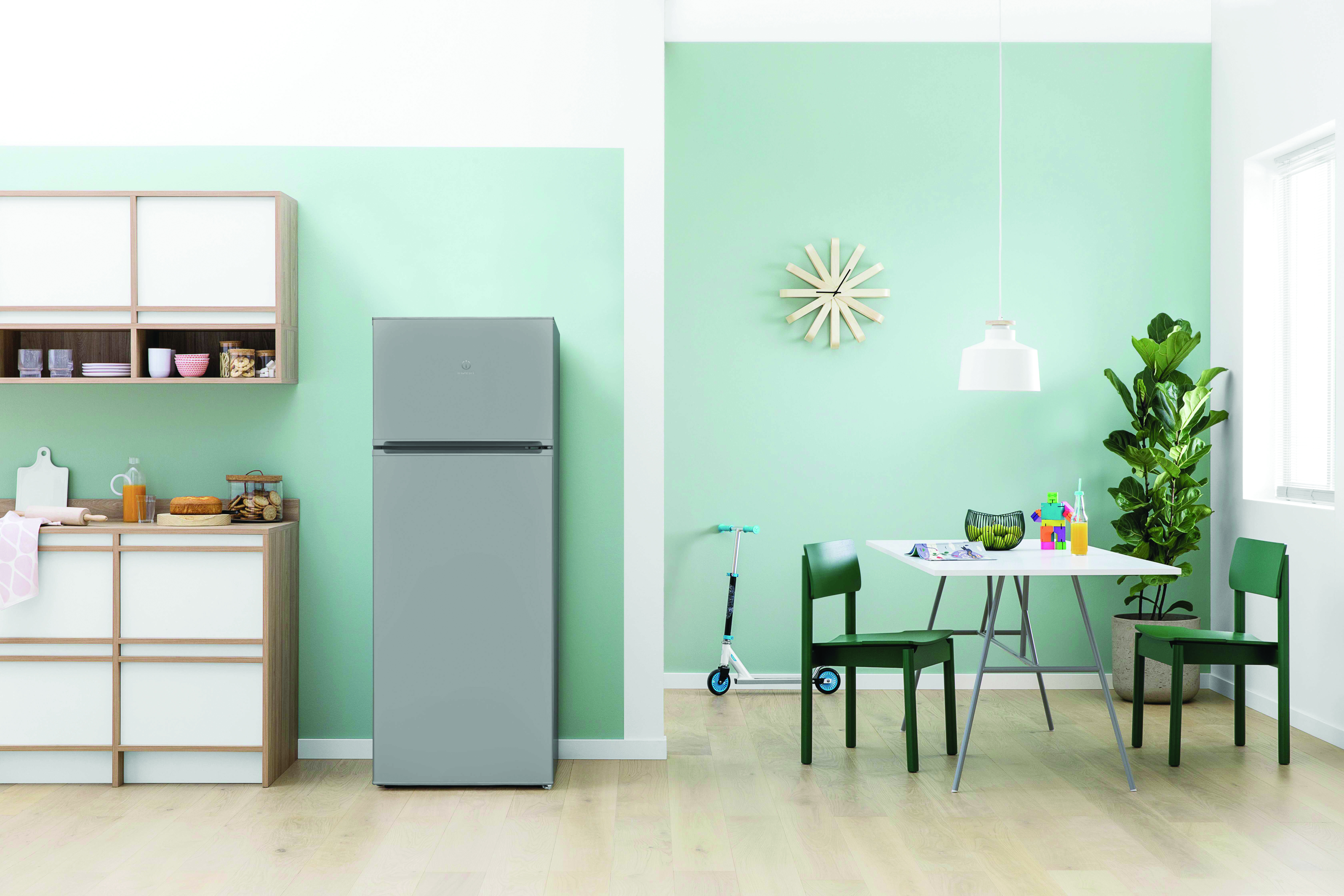 Una cocina con refrigerador, gabinetes y una mesa. También se ven un scooter y un reloj.
