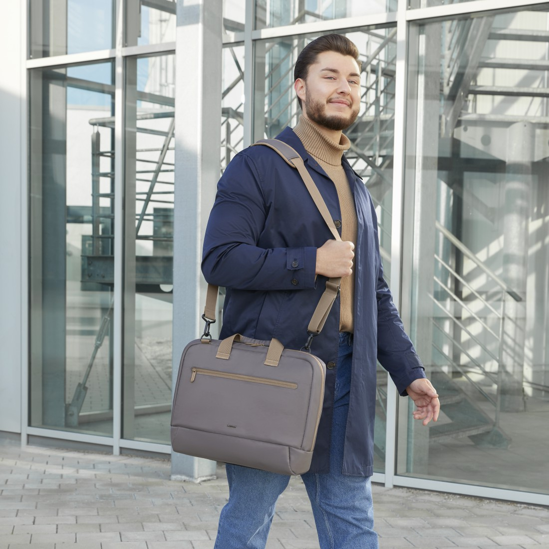Mann geht, trägt eine Laptoptasche. Glasgebäude im Hintergrund. Blauer Mantel, Jeans und Rollkragenpullover.