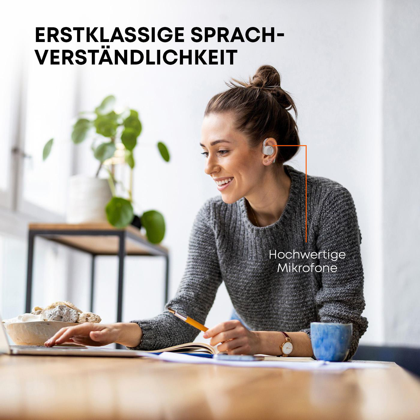 Eine Frau mit Kopfhörern lächelt, während sie an einem Laptop arbeitet. Ein Bleistift und ein Notizbuch liegen auf dem Tisch.