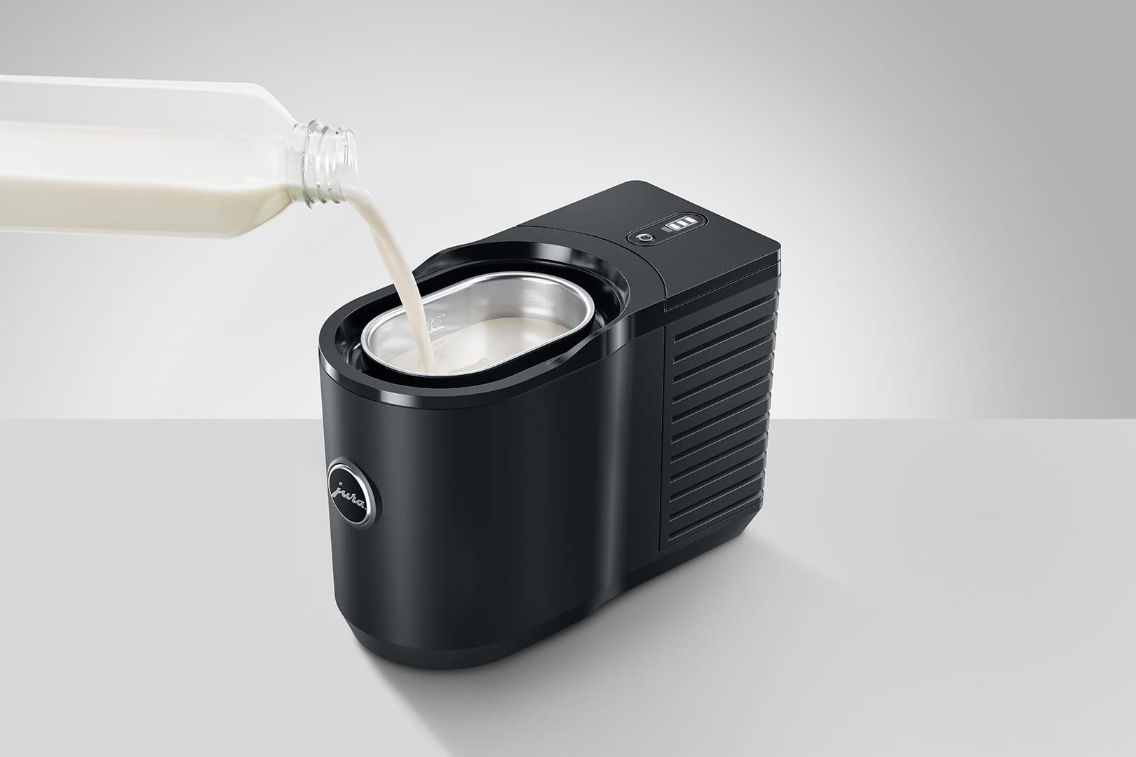 Milch wird in einen schwarzen Milchaufschäumer gegossen. Der Aufschäumer steht auf einer weißen Oberfläche.