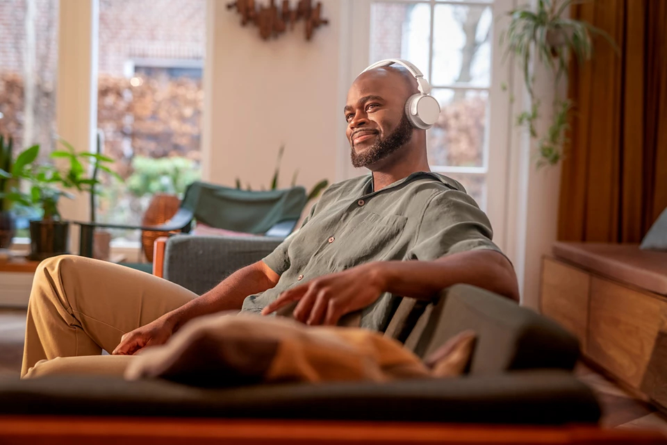 Mężczyzna w słuchawkach siedzi na kanapie, uśmiechając się. Pies leży przed nim na kanapie.