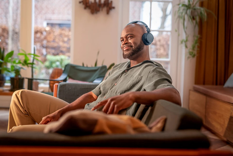 Mężczyzna ze słuchawkami siedzi na sofie, uśmiecha się i patrzy w dal, słuchając muzyki. Pies leży na sofie na pierwszym planie.