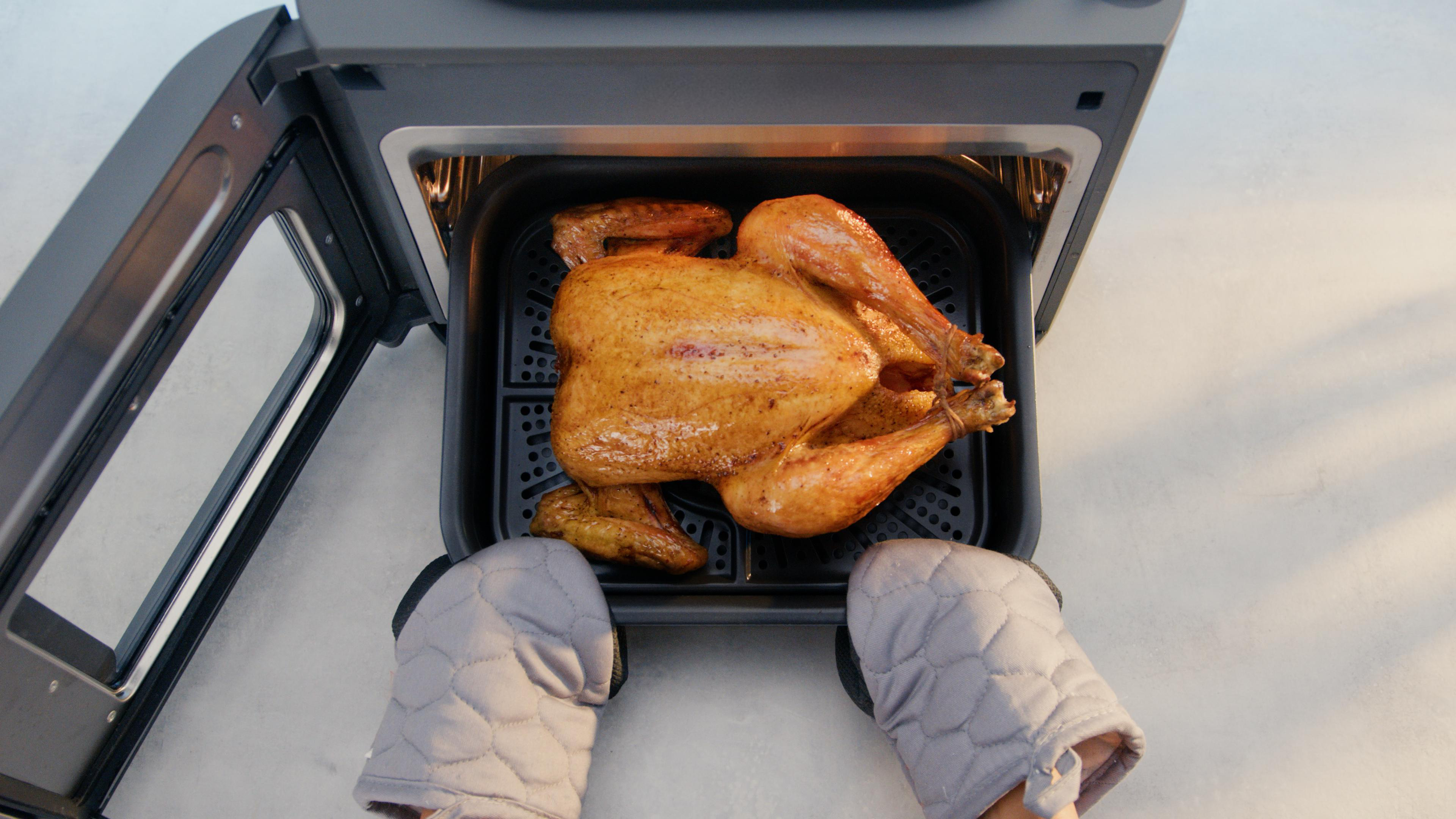 Ein gebratenes Huhn in einer Heißluftfritteuse, darunter Ofenhandschuhe. Die Tür ist geöffnet.