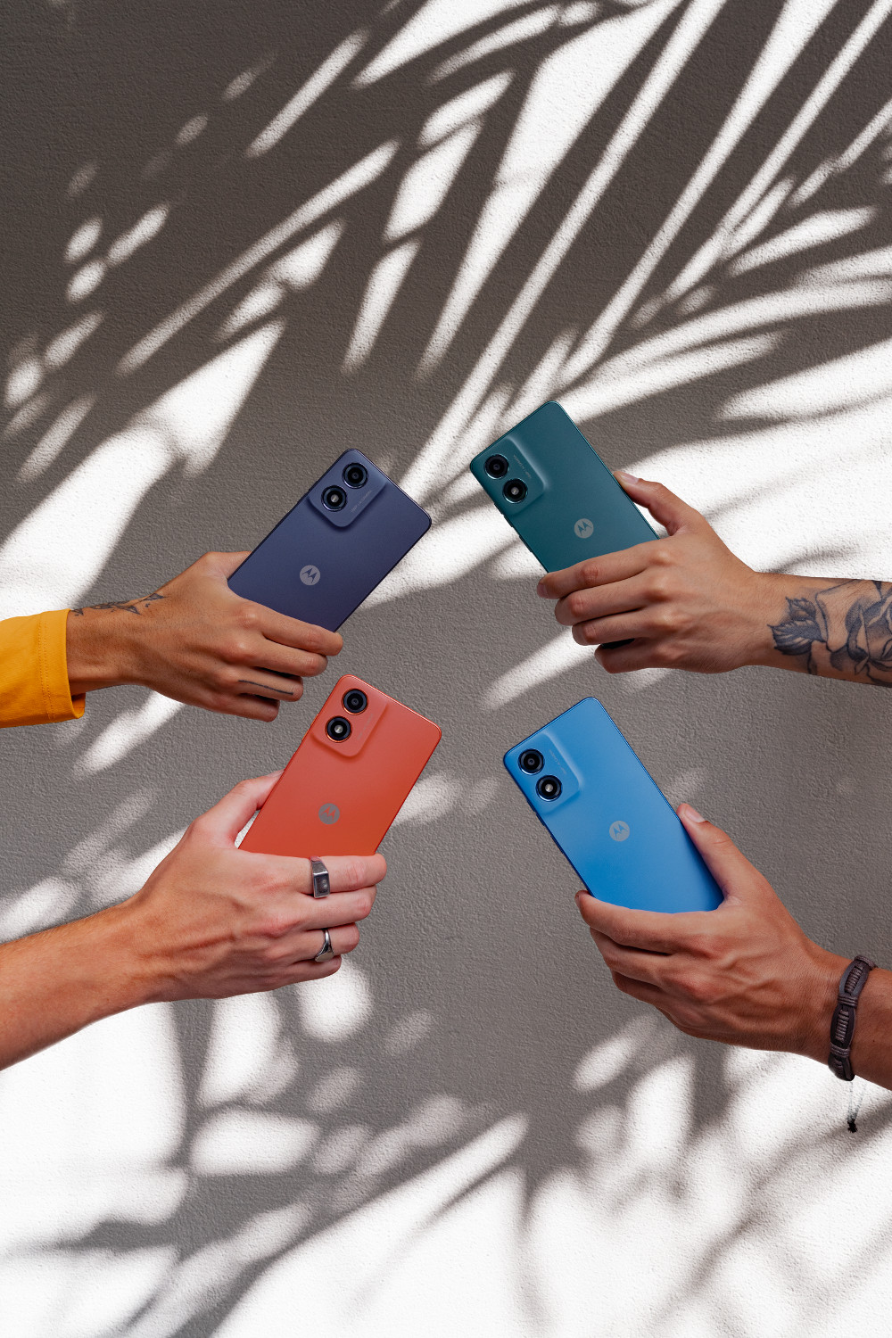 Cztery dłonie trzymające smartfony w różnych kolorach: niebieski, zielony, pomarańczowy i fioletowy. Cień liści na ścianie.