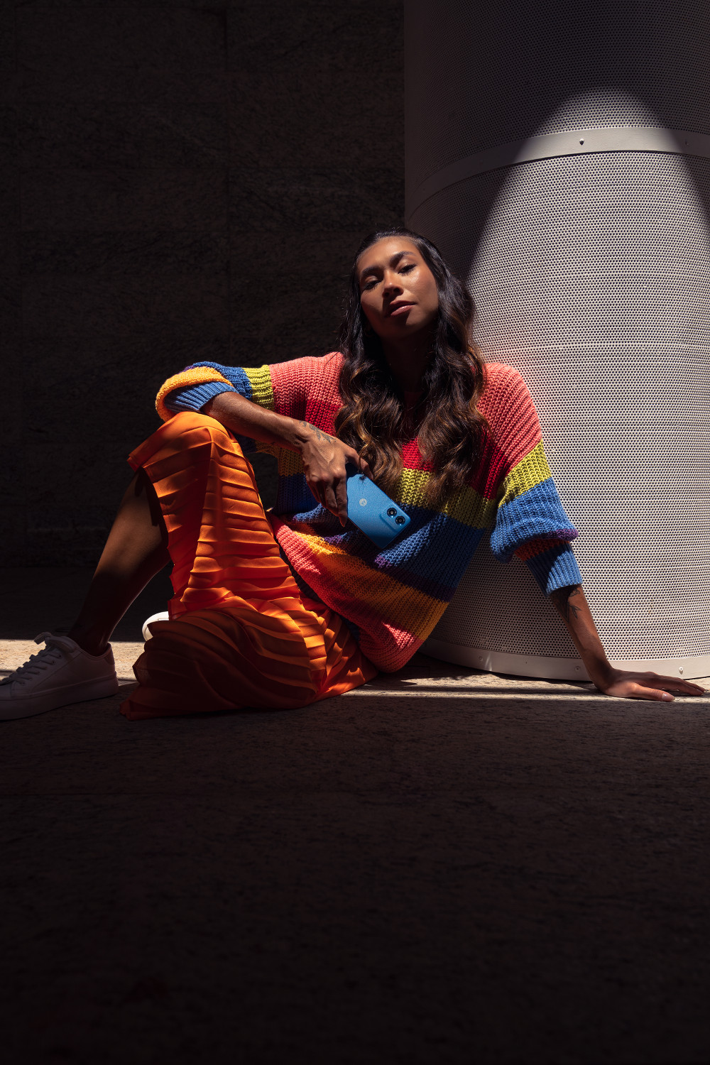 Kobieta siedzi, trzymając niebieski telefon. Ma na sobie pasiasty sweter i pomarańczową spódnicę. Biały głośnik w tle.