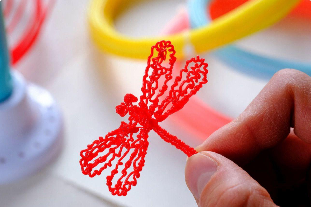 Rote Libellen-Skulptur in der Hand, bunte Drähte im Hintergrund.