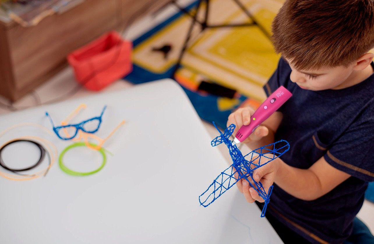 Junge, der einen rosa 3D-Stift verwendet, um ein blaues Flugzeug auf einer weißen Oberfläche zu erstellen.