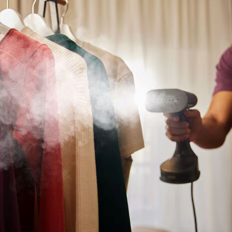 Osoba parująca ubrania parownicą ręczną, kilka koszul na wieszaku.