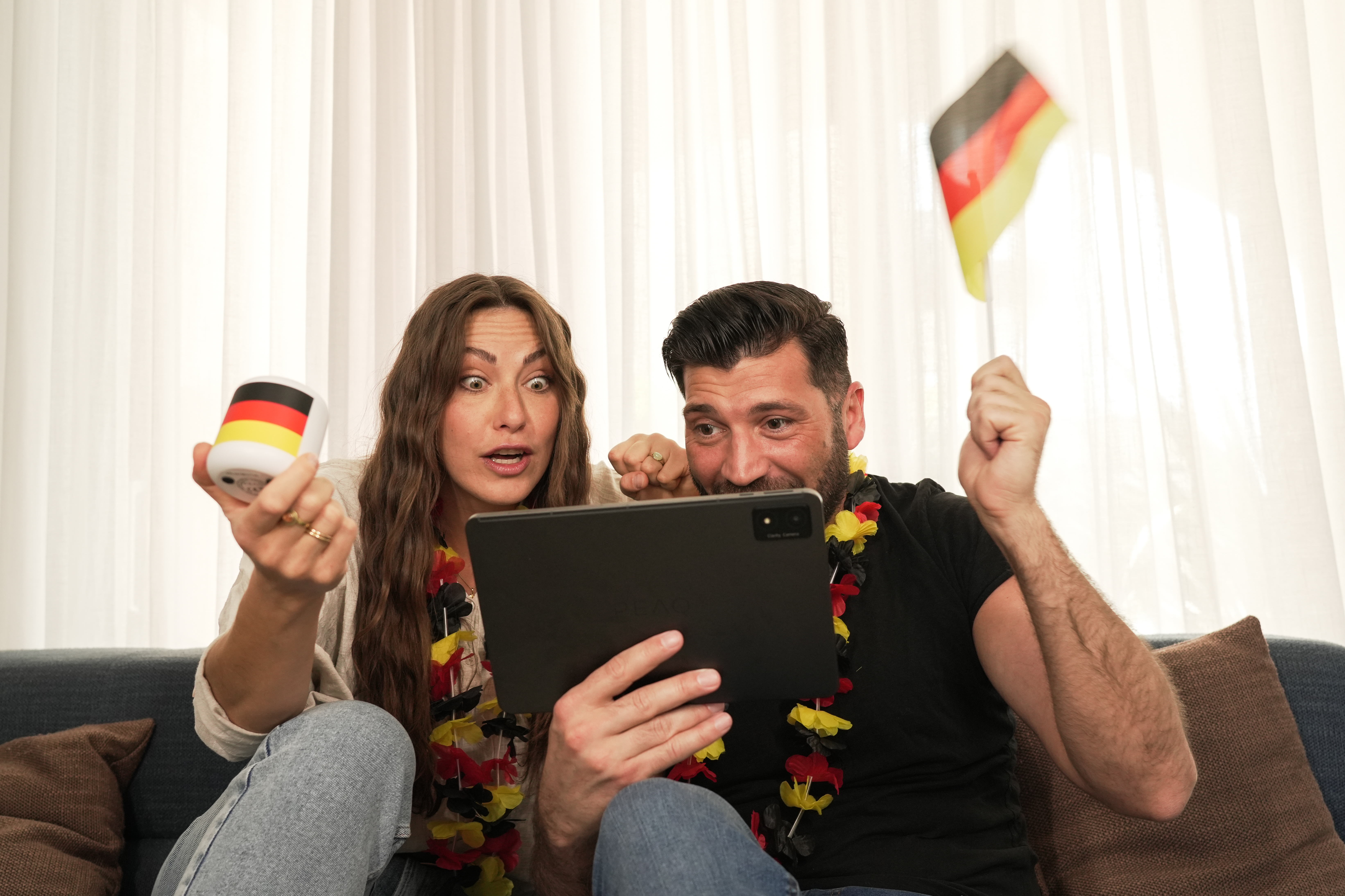 Zwei Personen schauen Tablet, halten Deutschlandfahne & farbigen Gegenstand, aufgeregte Gesichter.