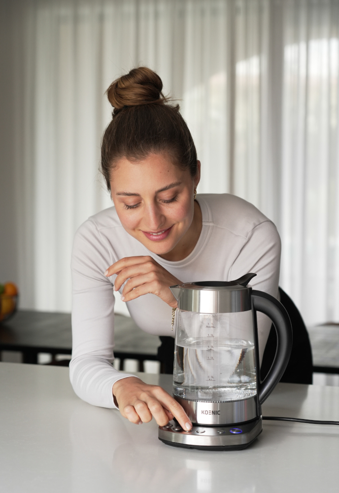 A woman looks at a glass electric kettle on a white counter, pressing a button.