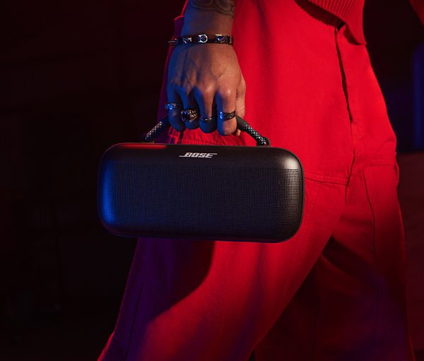 Osoba trzymająca czarny głośnik z logo 'BOSE'. Ma na sobie czerwone spodnie i pierścionki.