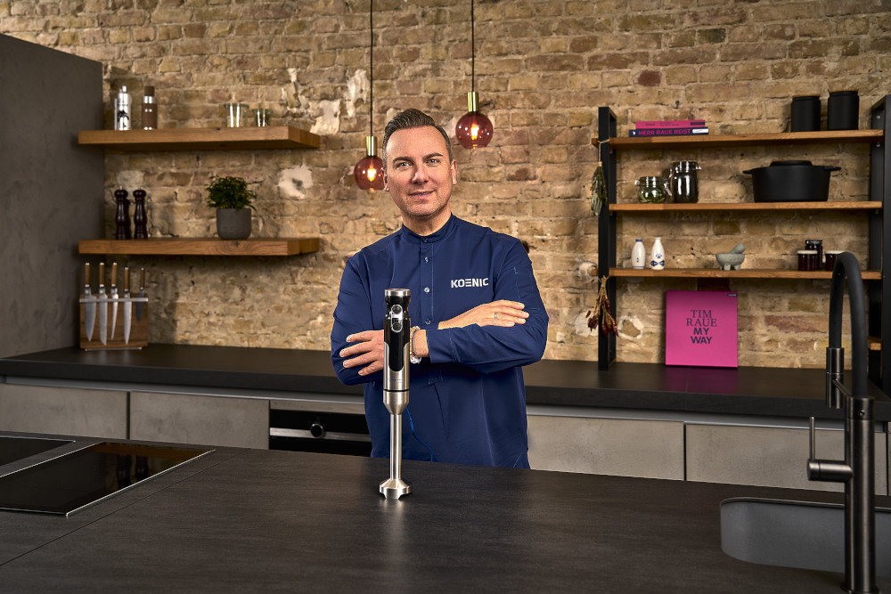 Man in een keuken met een zilveren en zwarte staafmixer, voor een aanrecht.