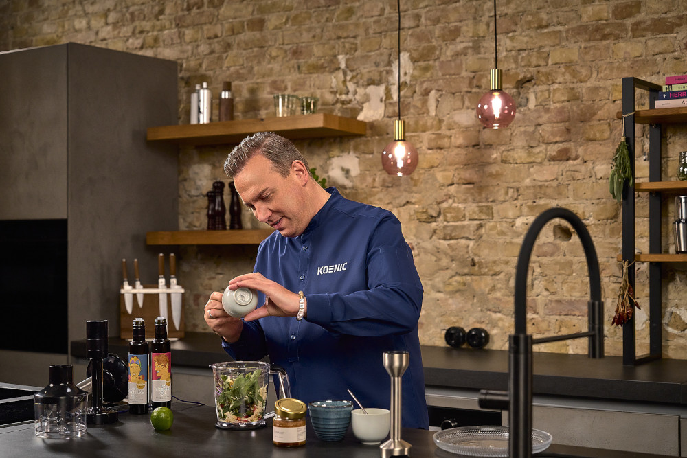 Chef-kok in blauw overhemd die ingrediënten toevoegt aan een blender in een keuken. Andere ingrediënten zijn zichtbaar.