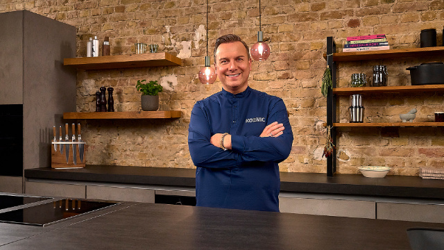 Een man in een blauw overhemd staat in de keuken met gekruiste armen. Schappen en een aanrechtblad erachter.