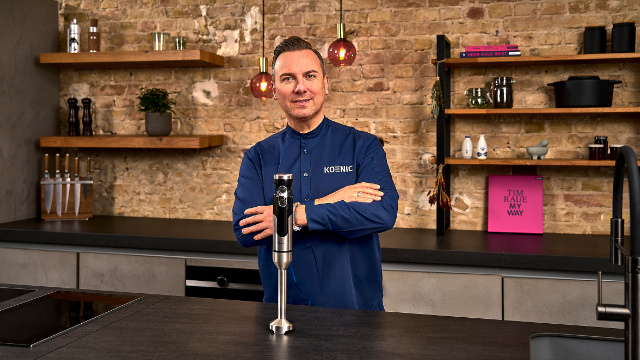 Een man in een blauw overhemd houdt een zilveren staafmixer vast in een keuken.