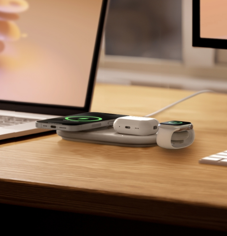 Egy iPhone, AirPods és Apple Watch töltődik egy fa íróasztalon egy laptop és monitor mellett.