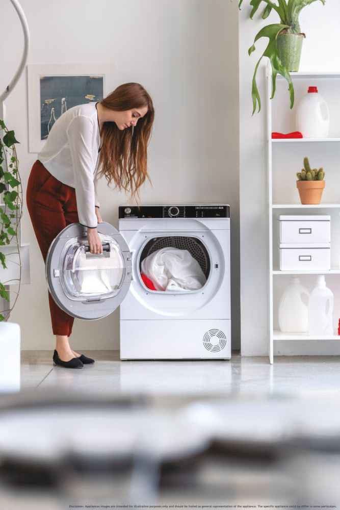 Femme mettant des vêtements dans un sèche-linge blanc. Porte ouverte, linge à l'intérieur. Buanderie.