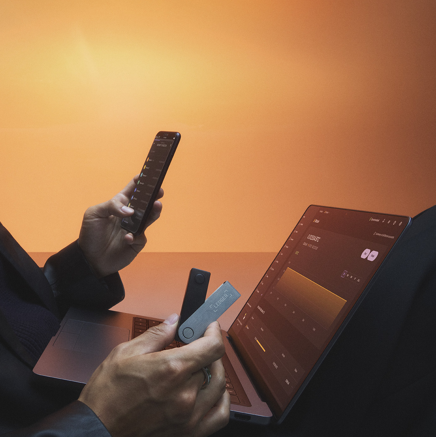 Bir kişi telefon ve iki Ledger donanım cüzdanını bir dizüstü bilgisayarın yanında tutuyor. Turuncu arka plan.