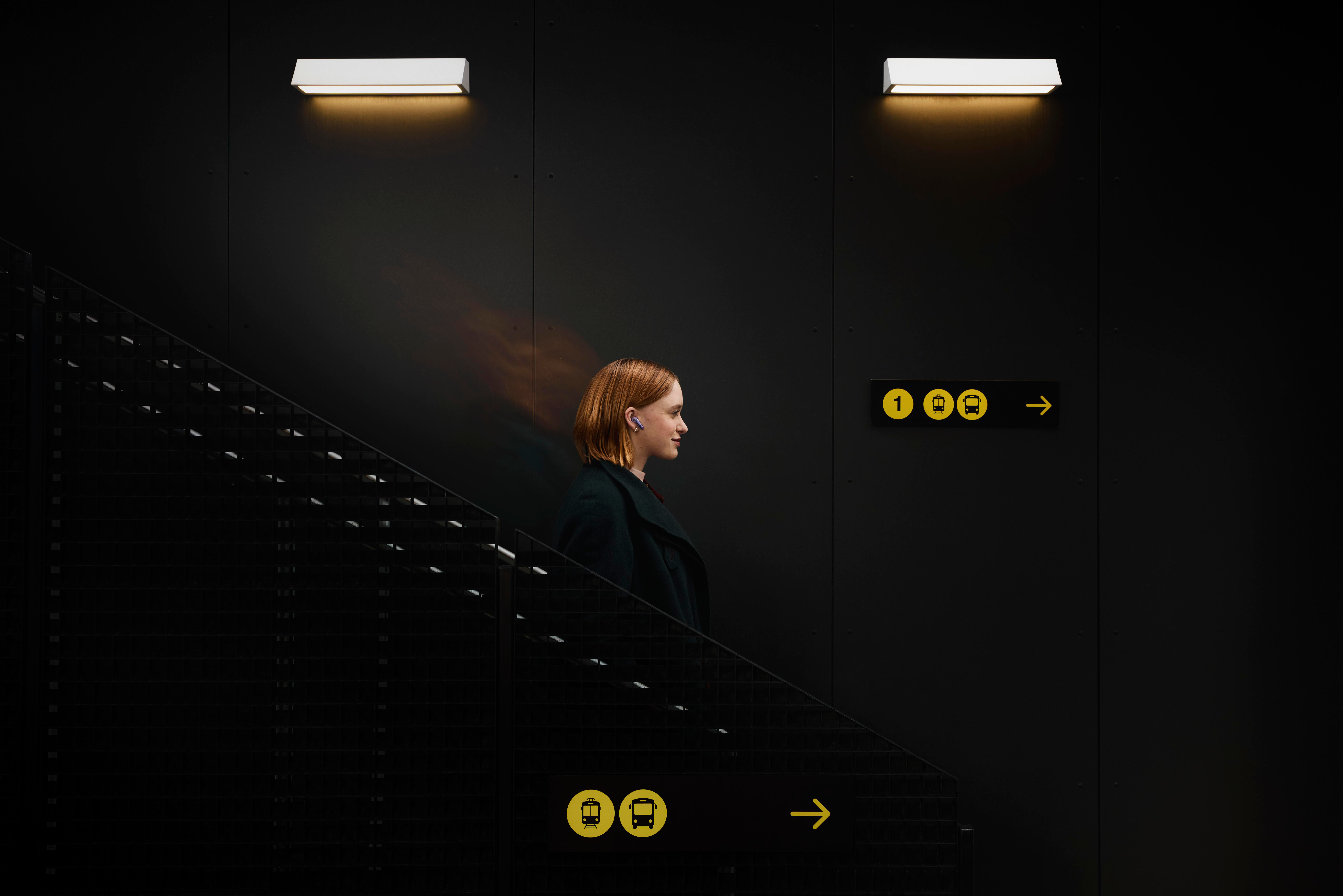 Eine Frau mit roten Haaren geht eine Treppe hinauf, Seitenansicht. Dunkler Hintergrund mit Lichtern und einem Schild.
