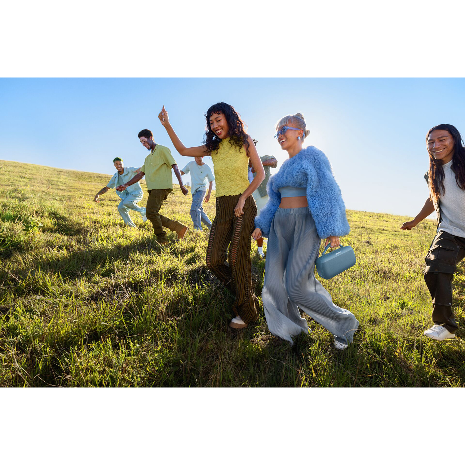 Gruppo di persone che corrono e ballano su una collina verde, con un altoparlante blu in primo piano.