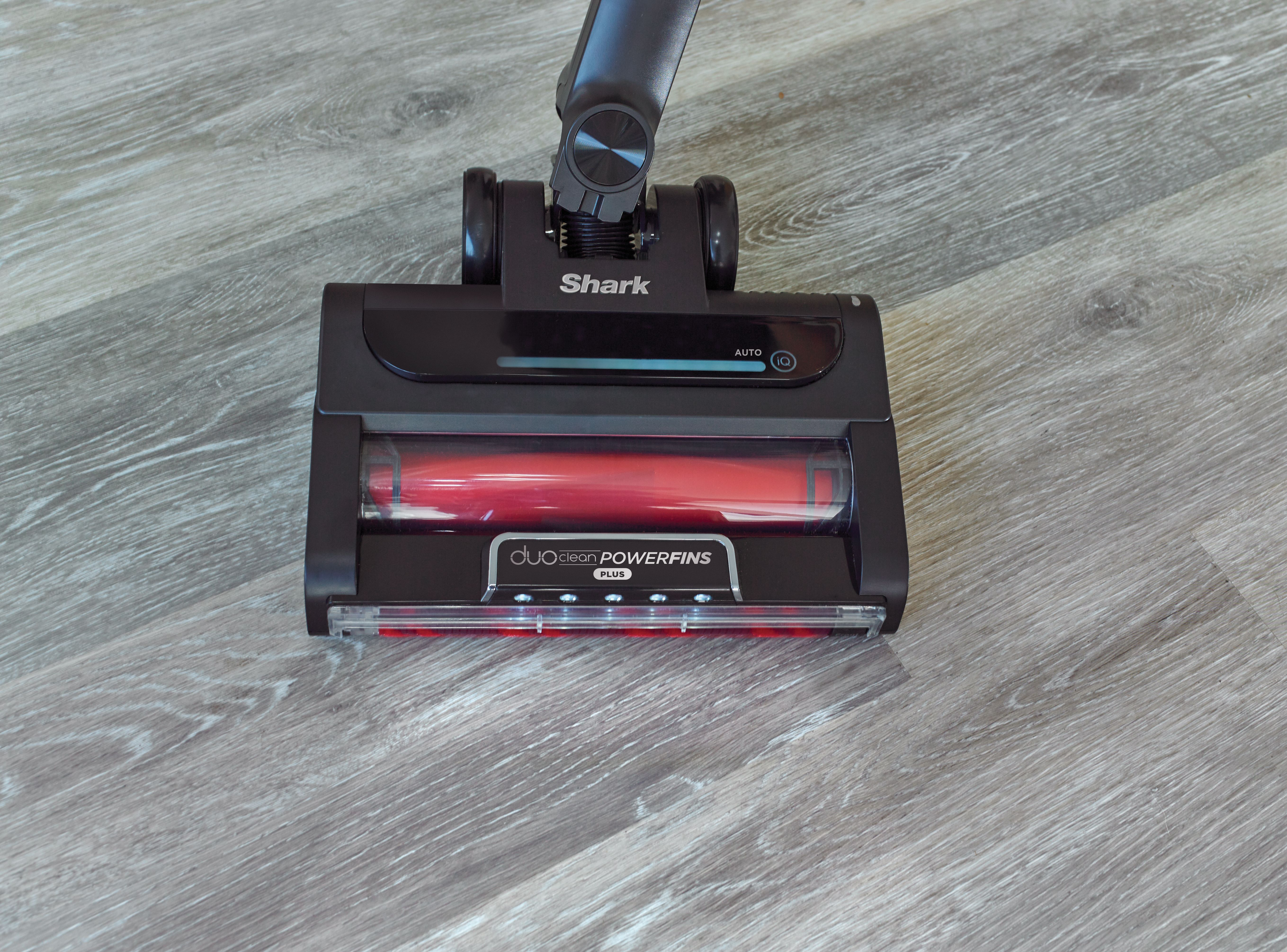Tête d'aspirateur noire sur sol en bois gris. Rouleau de brosse rouge visible. Logo Shark visible.
