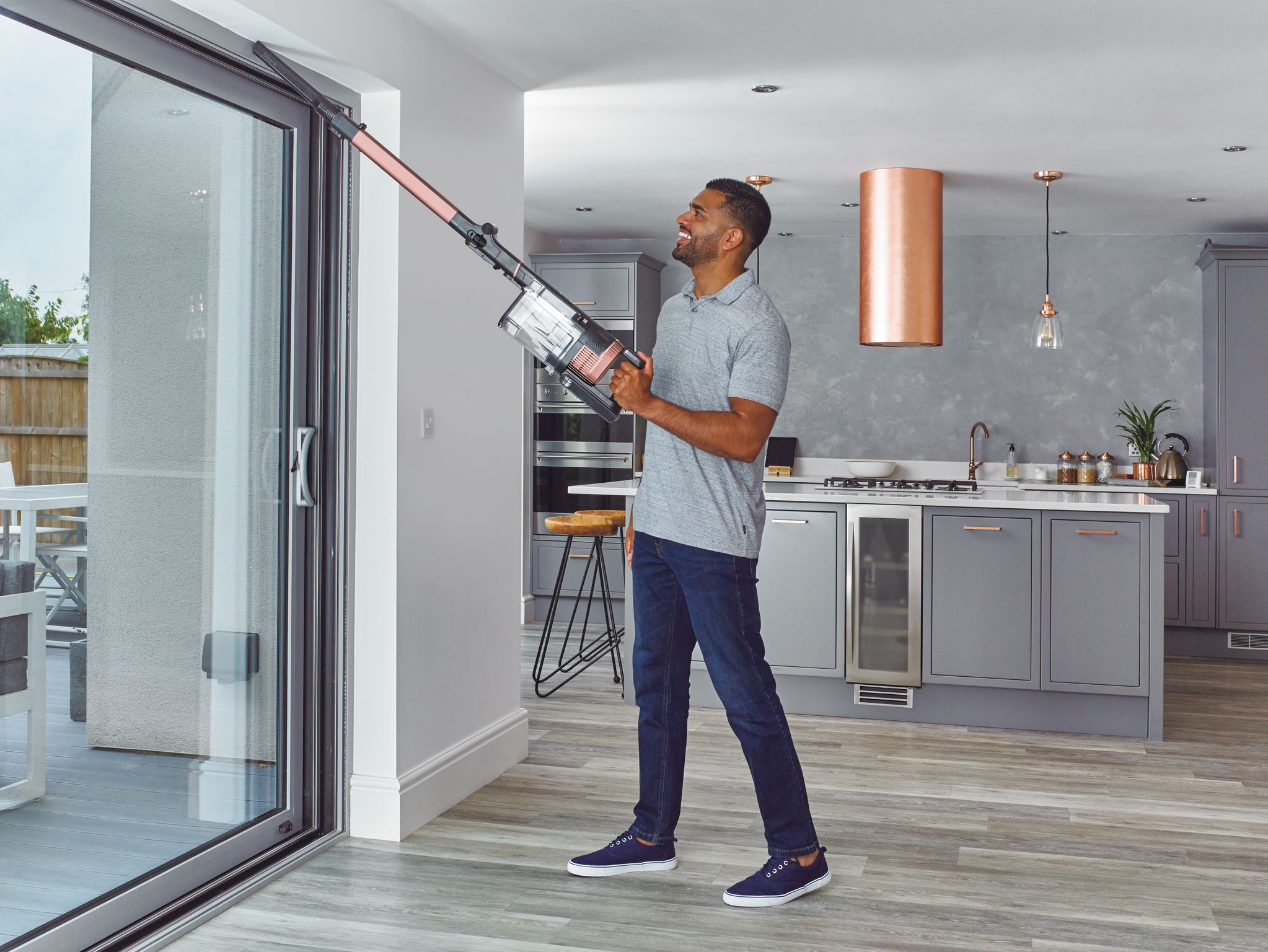 Homme passant l'aspirateur près d'une porte vitrée. Il sourit. La pièce a des murs clairs et une cuisine.