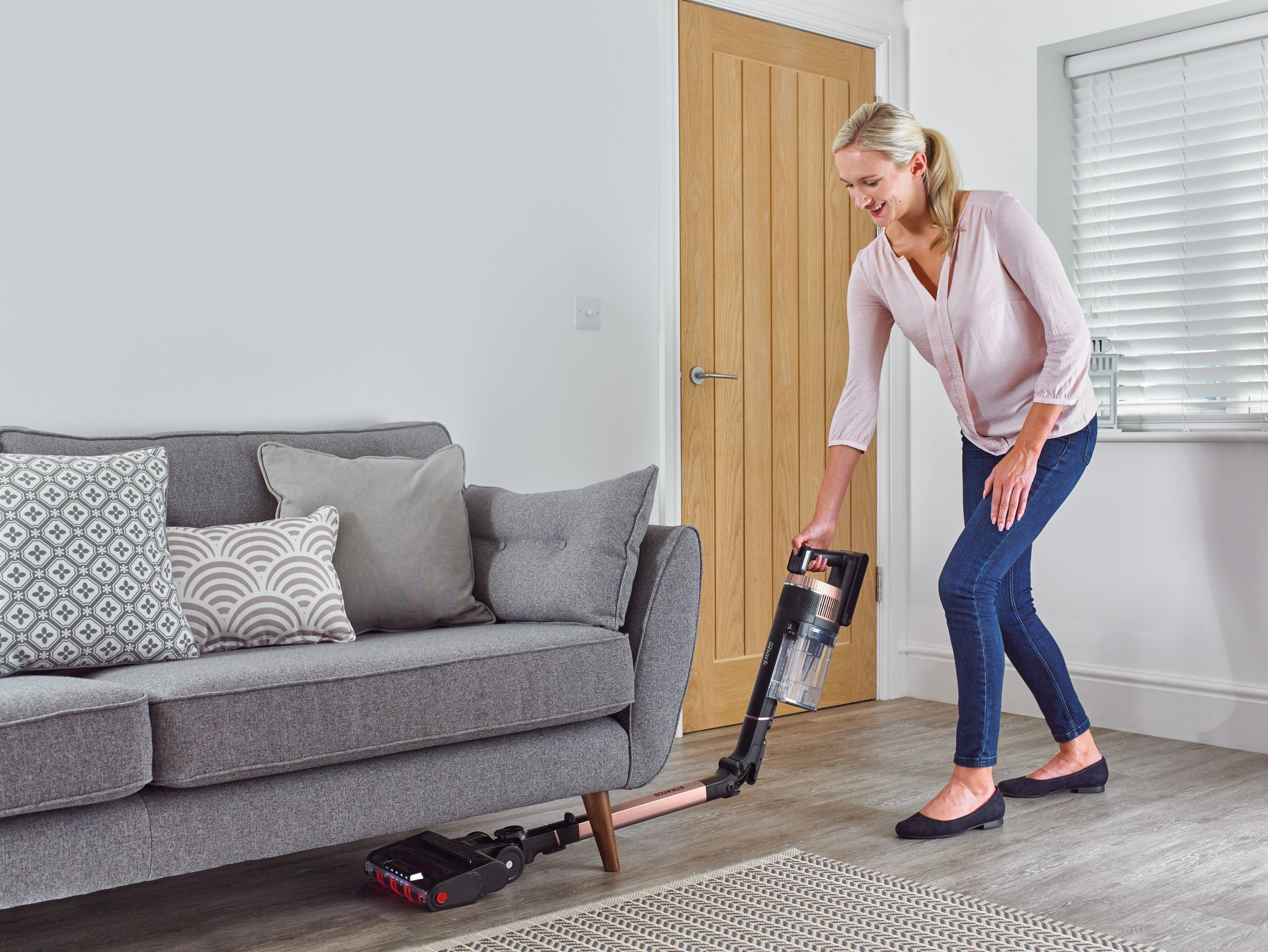 Une femme passe l'aspirateur dans un salon avec un canapé gris, une porte en bois et des planchers de bois franc.