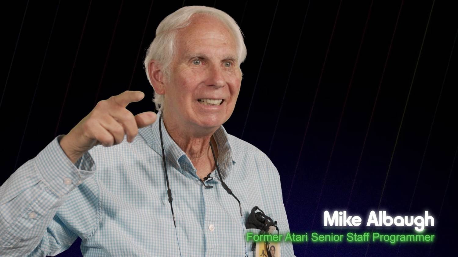 Hombre mayor sonriendo, señalando. Texto: 'Mike Albaugh, Former Atari Senior Staff Programmer' en barra verde.