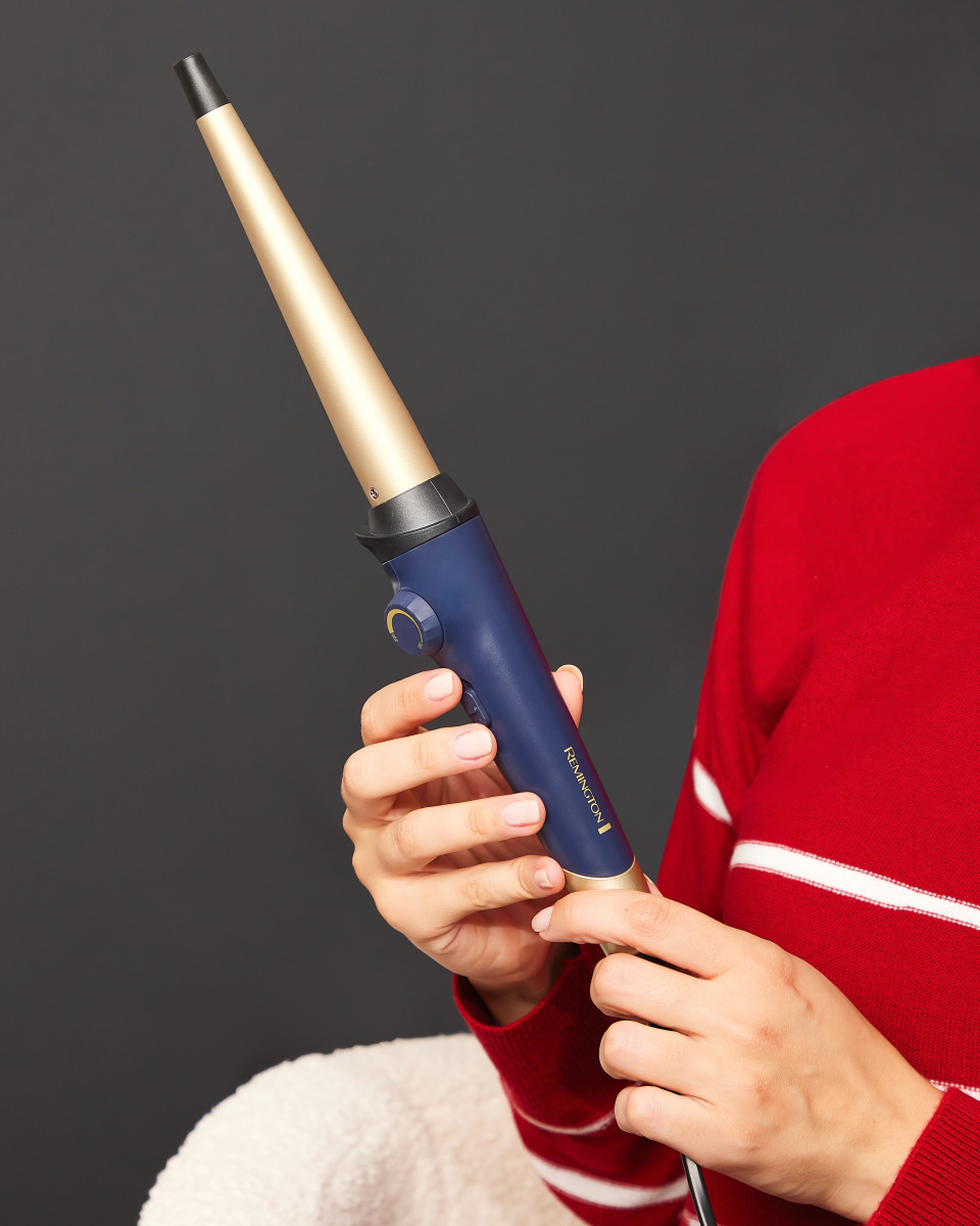 Una donna tiene in mano un arricciacapelli blu scuro e oro, con un maglione rosso.