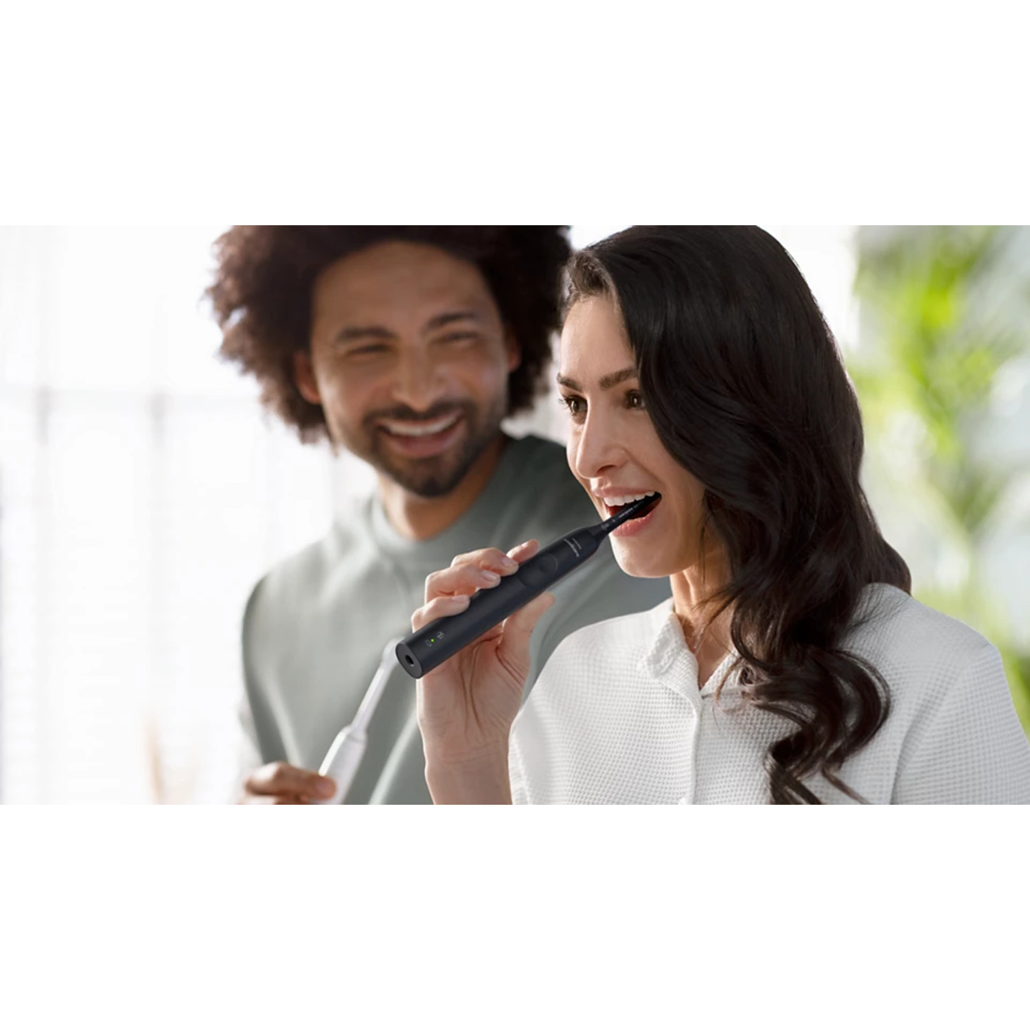 Femme se brossant les dents, souriante. Homme en arrière-plan souriant. Salle de bain blanche. Brosse noire.